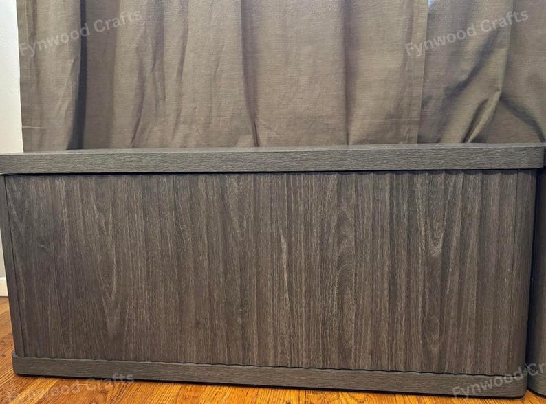 May include: A large, rectangular wooden storage chest with a dark gray wood grain finish. The chest has a flat top and a base, and is set against a neutral-colored curtain backdrop. The chest is designed for storage.
