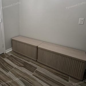 May include: Two light-colored wooden storage benches with a ribbed design on the front panel. The benches are rectangular with flat tops and are placed side-by-side against a light gray wall. The floor is tiled with a brown and gray pattern.