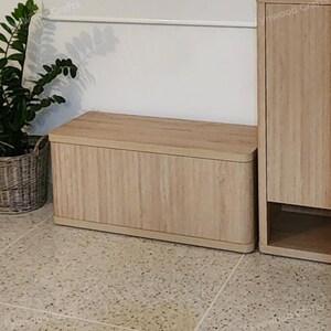 May include: A light-colored wooden storage bench with a rectangular shape and rounded edges. The bench is next to a matching wooden cabinet. A potted plant is in the background.