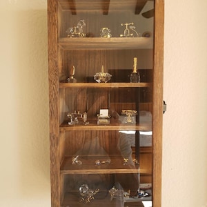 May include: A wooden display cabinet with a glass door, showcasing miniature glass figurines on multiple shelves. The cabinet has a warm brown finish and is mounted on a white wall. The figurines include animals, a typewriter, and a telephone.