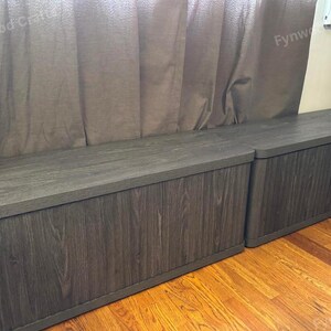May include: Two dark gray wooden storage benches with a wood grain finish. The benches have rounded corners and are placed on a hardwood floor. The background includes a brown curtain.