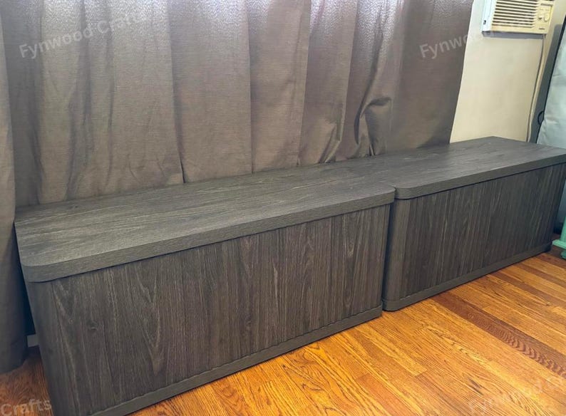 May include: Two dark gray wooden storage benches with a wood grain finish. The benches have rounded corners and are placed on a hardwood floor. The background includes a brown curtain.