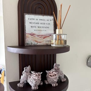May include: A dark brown wooden wall shelf with two semi-circular shelves. The top shelf holds a sign with the text "FAITH AS SMALL AS A MUSTARD SEED CAN MOVE MOUNTAINS". A glass diffuser and two decorative lion statues are on the shelves.