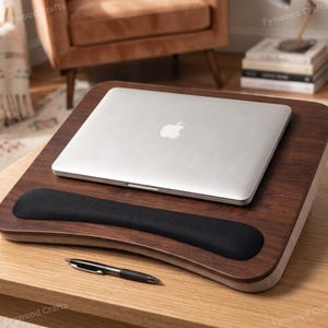 May include: A wooden laptop desk with a built-in black wrist rest. A silver laptop is placed on the desk. A black pen rests on the wooden surface. The desk is on a light-colored wooden table.