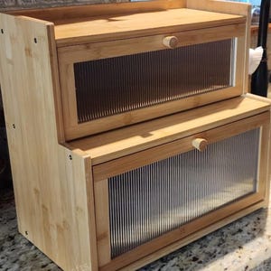 May include: A two-tiered bamboo bread box with clear, ribbed glass doors and wooden knobs. The natural wood grain is visible, and the box is designed to sit on a countertop. The overall design is simple and functional.