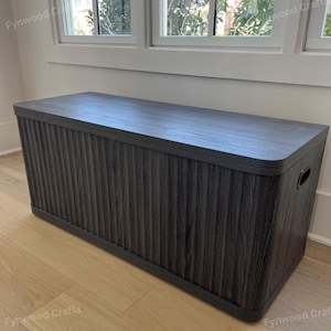 May include: A dark gray wooden storage chest with a ribbed front panel and a flat top. The chest has rounded corners and a handle on the side. It is placed against a white wall near a window.