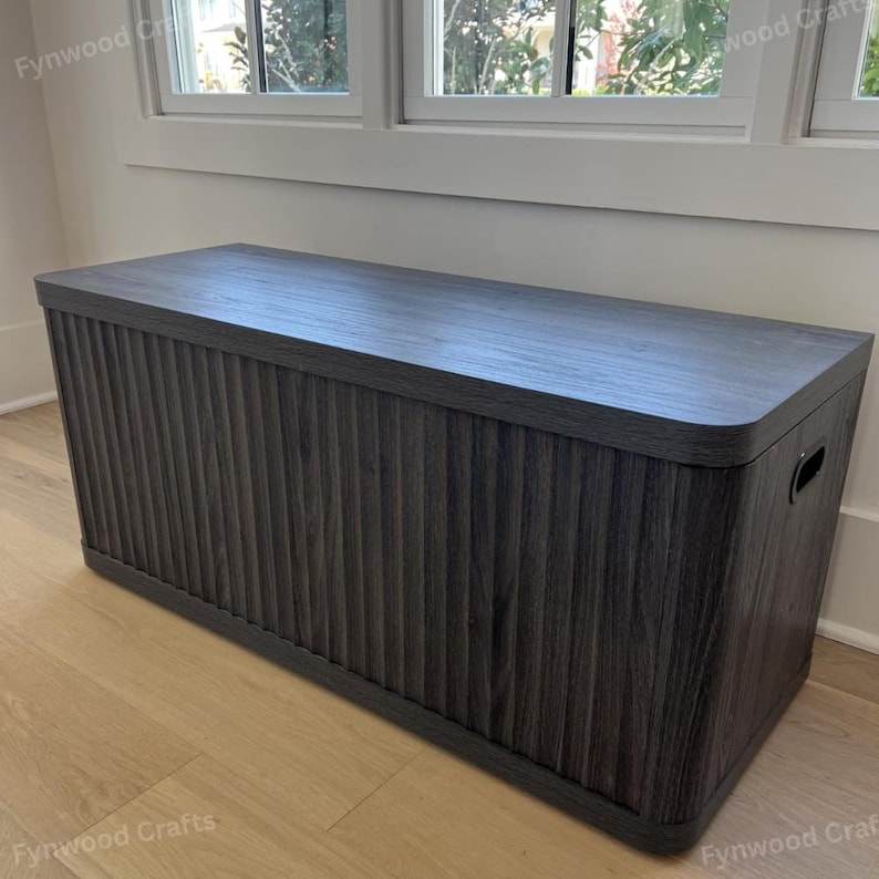 May include: A dark gray wooden storage chest with a ribbed front panel and a flat top. The chest has rounded corners and a handle on the side. It is placed against a white wall near a window.