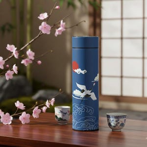 May include: A blue insulated tumbler with a Japanese crane design, featuring a red sun, white clouds, and waves. Two small ceramic teacups with floral patterns sit on a wooden table, alongside a branch of pink cherry blossoms.