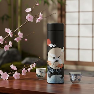 May include: Black insulated tumbler with a crane and floral design. The tumbler features a black lid and a decorative illustration of a crane, sun, and clouds. Two small teacups with blue and white patterns are also visible.