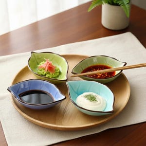 Könnte beinhalten: Ein hölzernes Serviertablett mit vier blattförmigen Keramikschalen in Grün, Blau und Türkis. Jede Schale enthält eine andere Zutat, darunter Wasabi, Sojasauce und eine cremige Sauce. Essstäbchen liegen auf einer Schale.