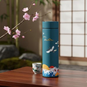 May include: A teal insulated water bottle with a Japanese-inspired design featuring cranes, clouds, and waves. A small teacup with a similar design sits nearby. The bottle is cylindrical and appears to be made of stainless steel.