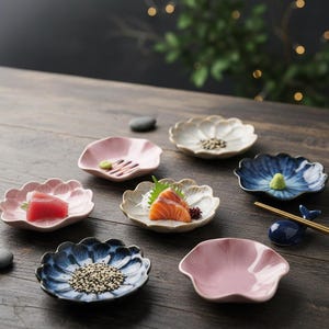 Puede incluir: Una colección de pequeños platos de cerámica con forma de flor en varios colores, incluyendo rosa, blanco y azul. Algunos platos contienen sushi, wasabi y semillas de sésamo. También hay un reposa palillos azul con forma de ballena.