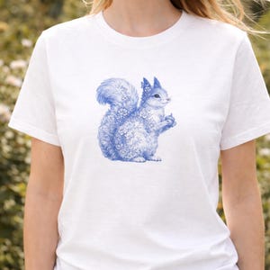 Puede incluir: Camiseta blanca con una ilustración azul detallada de una ardilla sosteniendo una nuez. La ardilla está adornada con detalles florales. El diseño está centrado en la parte delantera de la camiseta.