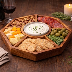May include: A wooden serving tray with multiple compartments, filled with snacks like cheese, crackers, olives, nuts, and salami. A small bowl of dip sits in the center. A lit candle, wine, and a bowl of blackberries are also present.