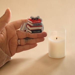 May include: A clear acrylic pin depicting a stack of books with the titles "Touch Her and Die", "Morally Gray", and "Villain". A red masquerade mask sits atop the books. A lit candle is nearby.