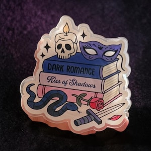 May include: A clear acrylic pin depicting a stack of books with the titles "Dark Romance" and "Kiss of Shadows." The pin also features a skull candle, a purple mask, a snake, a rose, a dagger, and a pair of red lips.