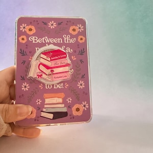 May include: A lilac phone case decorated with flowers and the words "Between the fa to be." The case features a central design of stacked books, with titles including "Enemies to Lovers" and "Forbidden Love".