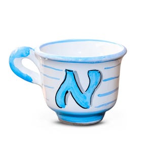 Può includere: Una tazza da tè in ceramica bianca con manico e bordo blu. La tazza presenta una grande lettera "N" blu stilizzata e strisce blu orizzontali. Anche la base della tazza è blu.