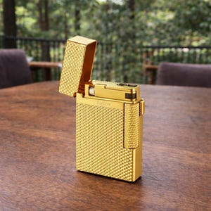 May include: A gold-coloured lighter with a textured, diamond-patterned surface. The top is open, revealing the interior mechanism. The word "Lighter" is printed on the front. The lighter is on a wooden surface.