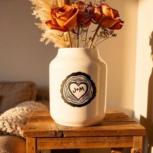 Op de afbeelding: Een witte keramische vaas met een zwart hartvormig ontwerp met de initialen "J+M" en een houtnerf rand. De vaas bevat oranje rozen en gedroogde siergrassen. De vaas staat op een rustieke houten tafel.