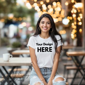 以下が含まれることがあります： 「Your Design HERE」の黒い文字が入った白いTシャツ。モデルはTシャツとブルージーンズを着用しています。背景は、ぼやけた屋外カフェの設定とストリングライトです。