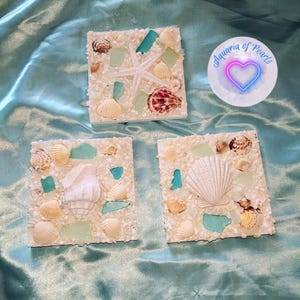Könnte beinhalten: Drei quadratische dekorative Untersetzer mit Strandmotiv. Jeder Untersetzer ist mit kleinen weißen Kieselsteinen bedeckt und mit Muscheln, Meerglas und einem Seestern verziert. Die Untersetzer sind auf einer hellblauen Satin-Oberfläche angeordnet.
