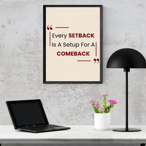 May include: A framed print with the quote "Every setback is a setup for a comeback" in red text. A black laptop, a small white flower pot with pink flowers, and a black desk lamp are on a white surface.