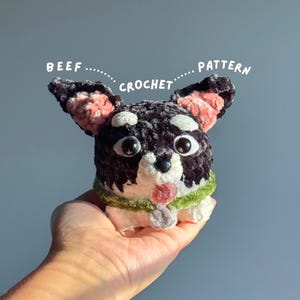 Pode incluir: Um brinquedo de pelúcia para cão feito em crochê com fios pretos, brancos e rosa, olhos de plástico grandes e uma coleira verde. O texto BEEF CROCHET PATTERN está escrito acima do brinquedo. O cão tem orelhas pontiagudas, nariz preto e língua rosa.