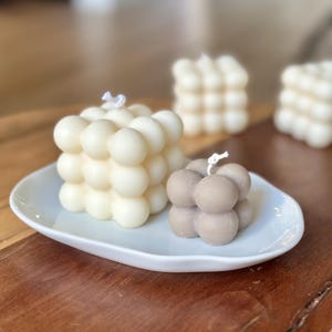 Peut inclure: Deux bougies à bulles, une ivoire et une taupe, sont posées sur un plat blanc de forme ovale. Les bougies ont un design sphérique. D'autres bougies à bulles sont visibles en arrière-plan. Un accessoire de décoration pour la maison.