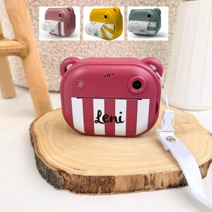 Puede incluir: Una cámara instantánea burdeos con un diseño de oso, con detalles a rayas blancas y burdeos y el nombre "Leni". La cámara tiene una lente, un flash y una correa blanca. Se muestran tres cámaras más pequeñas en amarillo, verde y burdeos arriba.