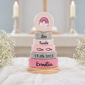 Könnte beinhalten: Ein stapelbares Holzspielzeug in Rosa- und Grautönen, gekrönt von einem Regenbogen-Design. Das Spielzeug trägt den Namen "Emilia" und das Datum "27.05.2023" sowie weitere Texte und Symbole. Es steht auf einem Holzsockel.