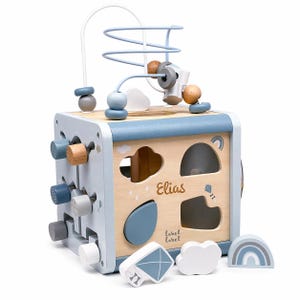 May include: A wooden activity cube in light blue and natural wood. It has a bead maze, shape sorter, and rotating blocks. The name "Elias" and the text "label label" are visible. Includes a kite, cloud, and rainbow.