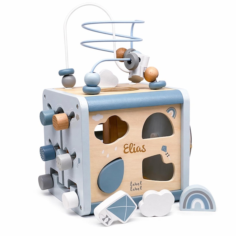 May include: A wooden activity cube in light blue and natural wood. It has a bead maze, shape sorter, and rotating blocks. The name "Elias" and the text "label label" are visible. Includes a kite, cloud, and rainbow.