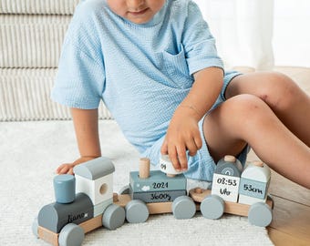 May include: A wooden toy train in shades of grey, white, and blue. The train features three carriages, each with unique details, including the name "Liano" and the numbers "2021" and "36005". The train is on a white rug.
