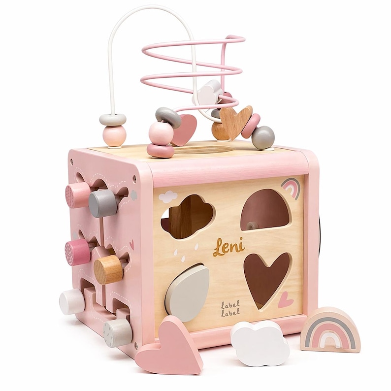 May include: A pink and natural wood activity cube with a bead maze, shape sorter, and other interactive elements. The cube features the name "Leni" and includes wooden shapes like hearts, clouds, and a rainbow. The design is child-friendly and colorful.
