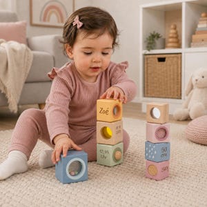 May include: A set of wooden sensory blocks in pastel colours, featuring cutouts with mirrors, beads, and printed text including the name Zoé, the date 01.10.2025, the time 12:45 Uhr, and measurements of 50 cm and 2500g, arranged in two vertical stacks.