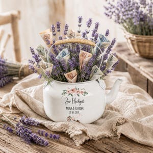 Peut inclure: Une théière en émail blanc remplie de brins de lavande violette et de billets en euros roulés. La théière présente un motif floral, le texte "Zur Hochzeit", "ANNA & FLORIAN" et la date "28.05.2026" imprimés avec une petite icône de clé.