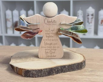 Puede incluir: Una figura de ángel de madera con las alas extendidas, sosteniendo billetes doblados. El ángel tiene una cabeza redonda y está montado sobre una rodaja de madera. El texto en el ángel dice "Charlotte 02.02.2025" y un mensaje en alemán.