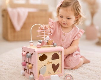 May include: A pink wooden activity cube with a bead maze and shape sorter. The cube features cut-out shapes, wooden pegs, and the name "Leni". Includes a small wooden rainbow and heart-shaped pieces. A fun toy for toddlers.