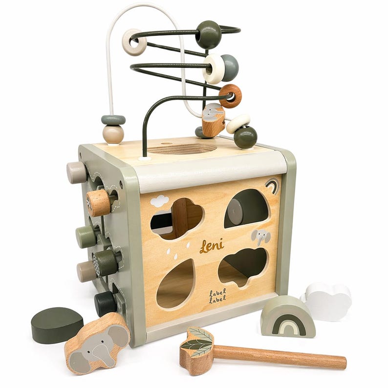 May include: A wooden activity cube featuring a bead maze, shape sorter, and pounding bench. The cube is painted in neutral tones of green, beige, and natural wood. Includes wooden shapes, a hammer, and the name "Leni".