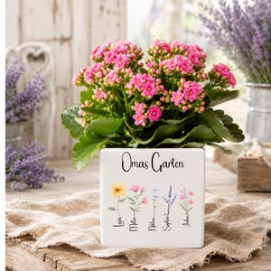 Puede incluir: Una maceta cuadrada de cerámica blanca que contiene una planta Kalanchoe de color rosa vibrante. El frente de la maceta presenta el texto Omas Garten sobre cinco flores silvestres ilustradas, cada una etiquetada con los nombres Leon, Milla, Niklas, Sophia y Julia.
