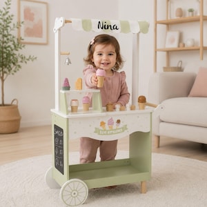 Può includere: Un carretto dei gelati in legno color verde salvia e bianco con una tenda a righe che riporta il nome Nina. Il carretto include una piccola campanella in metallo, una lavagna con i prezzi e vari coni e palline di gelato in legno in mostra.