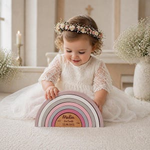 Puede incluir: Un juguete apilable de arcoíris de madera en colores pastel, incluyendo rosa, gris y blanco. El juguete tiene una base de madera personalizada con el nombre "Malia" y la fecha "23.05.2023". Una niña con un vestido blanco y una corona de flores juega con él.