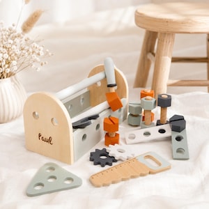 May include: Wooden toy toolbox with tools, including a hammer, saw, and screws. The toolbox is light brown with gray and orange accents. The name "Paul" is written on the side. The tools are arranged on a white surface.