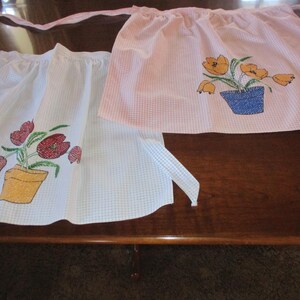 Puede incluir: Dos delantales con bordados florales. Uno es blanco con flores rojas en una maceta amarilla, el otro es rosa con flores naranjas en una maceta azul. Ambos delantales tienen un estampado de cuadros y lazos largos.