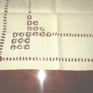 May include: Cream-colored linen tablecloth with intricate cutwork and decorative borders. The cutwork features geometric patterns and floral motifs, adding a touch of elegance to the textile. The tablecloth is folded, showing the detailed craftsmanship.