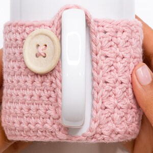 May include: A light pink crocheted mug cozy with a wooden button closure. The cozy is wrapped around a white mug, with the handle visible. The crochet stitch creates a textured surface. The button is round with four holes.