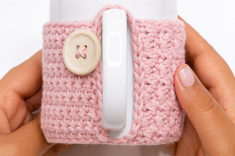 May include: A light pink crocheted mug cozy with a wooden button closure. The cozy is wrapped around a white mug, with the handle visible. The crochet stitch creates a textured surface. The button is round with four holes.