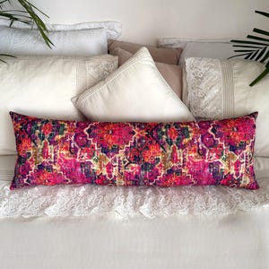 May include: A long, rectangular decorative pillow with a vibrant, patterned design in shades of pink, red, purple, and gold. The pillow is placed on a white bed with other pillows and a lace-trimmed bedspread.
