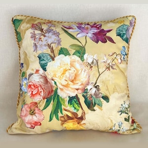 Könnte beinhalten: Quadratisches Zierkissen mit floralem Muster. Das Kissen hat einen gelben Hintergrund und ein farbenfrohes Blumenarrangement in Rosa, Pfirsich, Lila und Weiß. Die Ränder sind mit einer goldenen Kordel versehen.